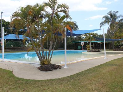Palm trees centre frame with the outdoor pool at Eagleby Aquatic Centre behind. Blue shade sail cover the pool left and right
